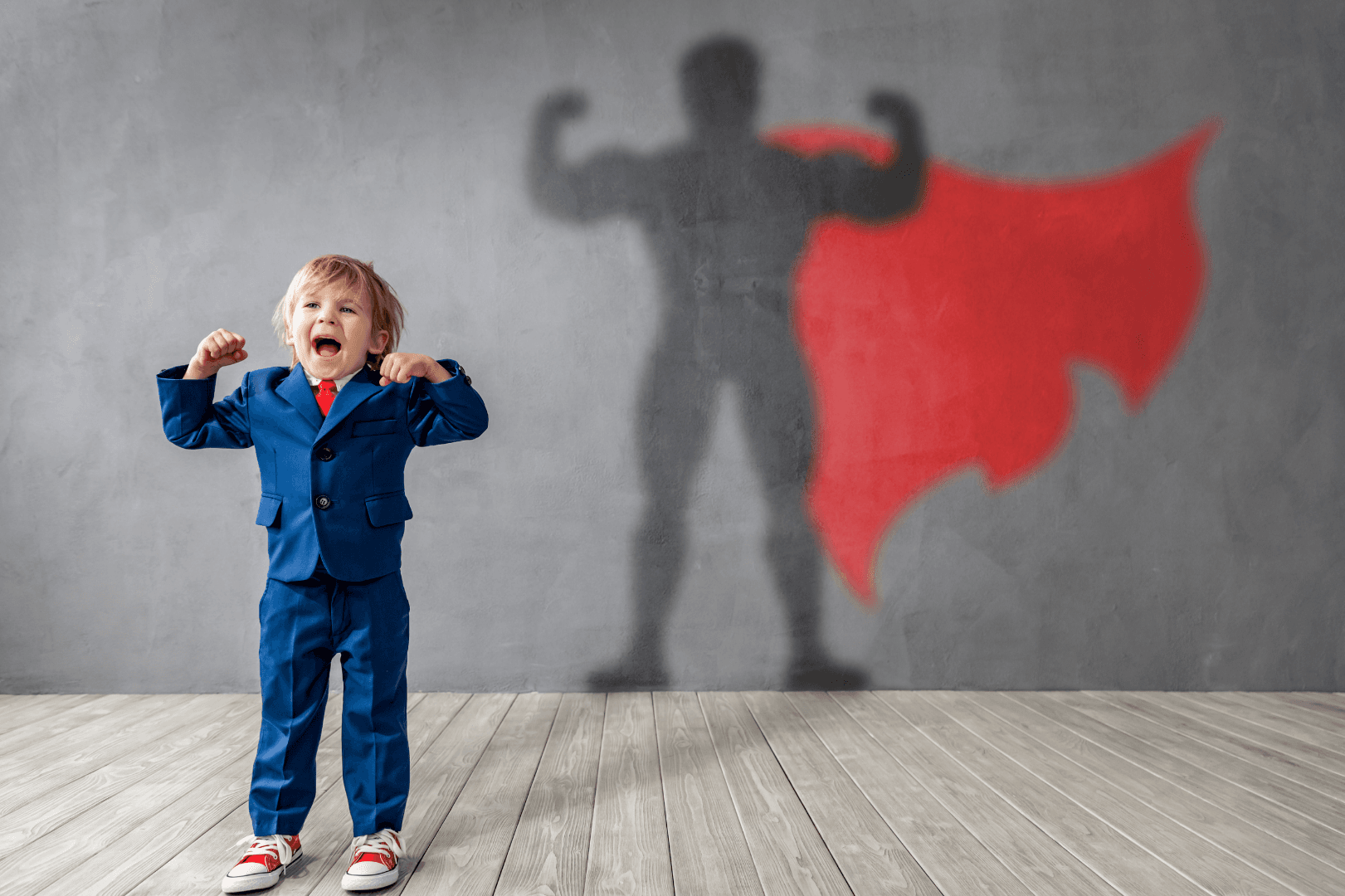 A child celebrating with arms raised; a superhero shadow with a red cape appears on the wall behind them.